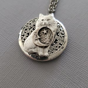 K&ouml;nnte beinhalten: Ein silberfarbener, runder Medaillon-Anh&auml;nger an einer passenden Kette. Das Medaillon zeigt ein erhabenes Relief einer Katze, die vor einer Mondsichel und einem Stern sitzt. Der Hintergrund des Medaillons hat ein komplexes, durchbrochenes Filigranmuster.