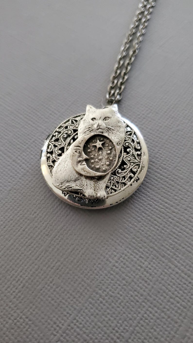 K&ouml;nnte beinhalten: Ein silberfarbener, runder Medaillon-Anh&auml;nger an einer passenden Kette. Das Medaillon zeigt ein erhabenes Relief einer Katze, die vor einer Mondsichel und einem Stern sitzt. Der Hintergrund des Medaillons hat ein komplexes, durchbrochenes Filigranmuster.