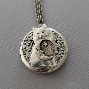 K&ouml;nnte beinhalten: Ein silberfarbener, runder Medaillon-Anh&auml;nger an einer passenden Kette. Das Medaillon zeigt ein erhabenes Relief einer Katze, die ein Mondsichel- und Sternmotiv vor einem Hintergrund aus filigranen, durchbrochenen Metalldetails h&auml;lt.