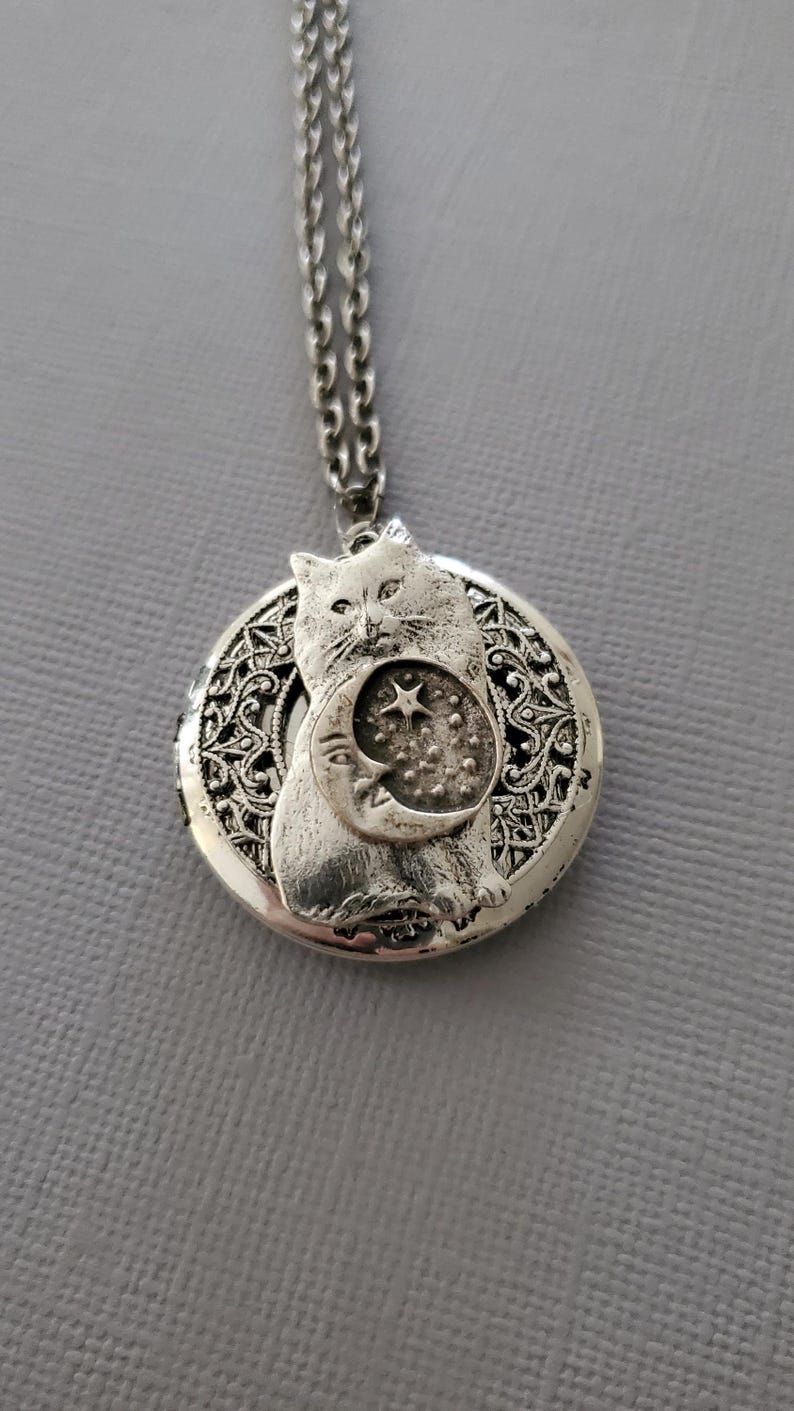 K&ouml;nnte beinhalten: Ein silberfarbener, runder Medaillon-Anh&auml;nger an einer passenden Kette. Das Medaillon zeigt ein erhabenes Relief einer Katze, die ein Mondsichel- und Sternmotiv vor einem Hintergrund aus filigranen, durchbrochenen Metalldetails h&auml;lt.