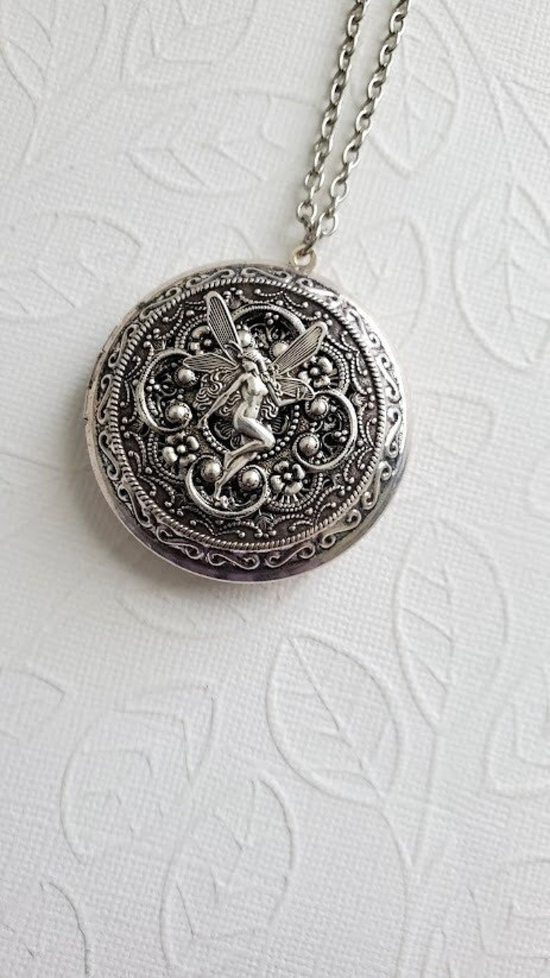 May include: Silver locket necklace with a detailed design featuring a fairy with wings. The locket has a filigree pattern and is on a silver chain.