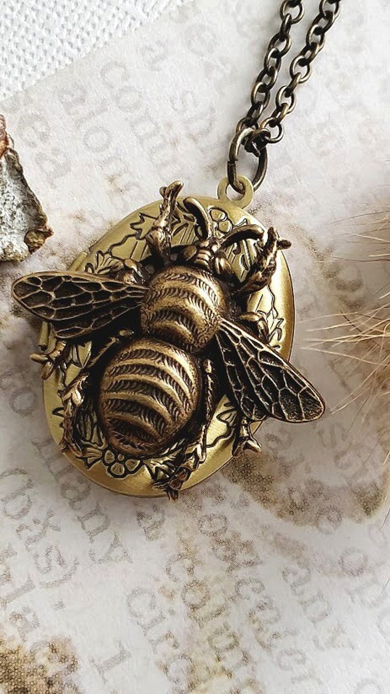 May include: A gold-toned necklace with a locket featuring a detailed bee design. The locket is oval-shaped and has a textured surface. The bee is depicted with intricate wings and a segmented body.