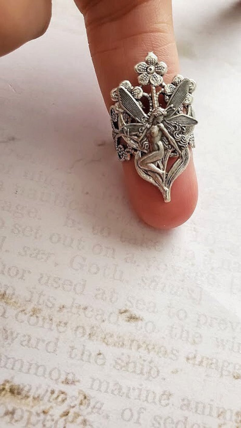 May include: Silver ring featuring a fairy with wings, sitting on a flower with leaves. The ring is detailed and intricate.