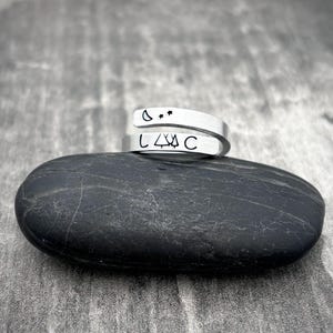 May include: Two silver wrap rings with stamped designs. One ring features a crescent moon and two stars, the other displays the letters "L4C" with a mountain symbol. The rings are displayed on a dark grey stone.