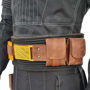 May include: A brown leather utility belt featuring a gold-colored rectangular buckle with geometric line detailing, two attached brown leather pouches, and a side section with yellow cylindrical components, worn over a black textured garment.