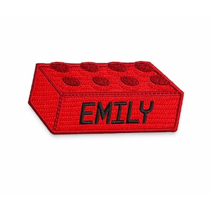 May include: A red embroidered patch in the shape of a building block. The block has eight raised studs on top and the name "EMILY" embroidered in black on the front. The patch is on a white background.