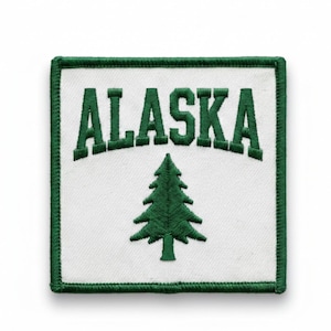 May include: Square patch with a white background, green border, and the word "ALASKA" embroidered in green block letters. A green pine tree is centered below the text. The patch is likely for a jacket or bag.