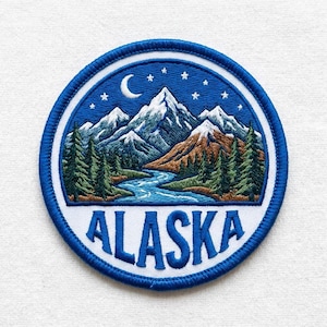 May include: Embroidered patch featuring a mountain landscape with a river, trees, and a crescent moon and stars against a blue sky. The word "ALASKA" is embroidered in white on a white banner. The patch has a blue border.