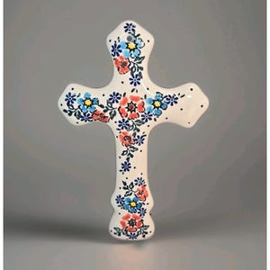 May include: A white ceramic cross wall hanging featuring a hand-painted floral pattern with red, light blue, and dark blue flowers, green leaves, and small black dots. The cross has a smooth, glossy finish and a small hole at the top for hanging.