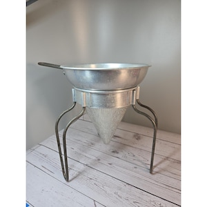 Könnte beinhalten: Eine silberfarbene Passiermühle oder ein Spitzsieb aus Metall mit einem breiten, runden oberen Becken, einem konischen Siebboden aus Metallgeflecht und drei geschwungenen Metallbeinen für Stabilität. Der Rahmen trägt die Prägung Foley Food Mill.