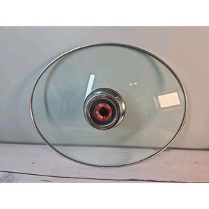 May include: A circular clear glass lid for cookware featuring a polished silver-tone metal rim and a central metallic knob. The knob includes a black centre with a red circular accent containing the white text Ultrex printed on the surface.