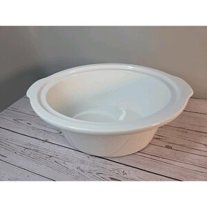 Può includere: Una ciotola in ceramica bianca da inserire in una pentola a cottura lenta, caratterizzata da una finitura liscia e lucida. Il recipiente rotondo presenta un bordo largo e piatto con manici laterali integrati per l'uso in una base.