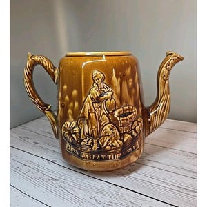 May include: Brown ceramic teapot with a detailed relief scene of a woman at a well. The teapot has a curved handle and spout, and the words "REBEKAH AT THE WELL" are inscribed on the front. The teapot is on a white wooden surface.