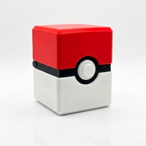 May include: A red and white cube-shaped container with a black band and a white circle with a black outline. The top is red, and the bottom is white. The container is a replica of a popular animated series item.