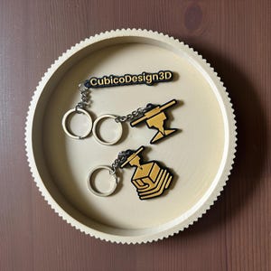 Pode incluir: Três porta-chaves com argolas e correntes de metal prateado. Um porta-chaves é uma etiqueta retangular preta com o texto amarelo CubicoDesign3D. Os outros dois apresentam designs de bocal e cubo de impressora 3D em amarelo e preto.