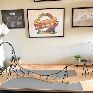 Pode incluir: Um modelo de montanha-russa com uma pista e suportes pretos, colocado sobre uma base de madeira. A montanha-russa está em uma mesa, com obras de arte emolduradas e uma lâmpada no fundo. A obra de arte inclui o texto "Coaster Highlight Z".
