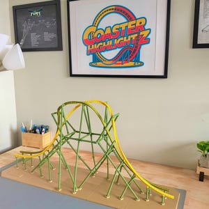 May include: A model roller coaster with a green frame and yellow track. The coaster is on a tan base. A framed print with the words "COASTER HIGHLIGHTZ" hangs on the wall behind the model.
