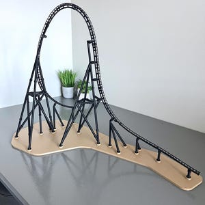 Puede incluir: Un modelo en miniatura de montaña rusa negra con un bucle y una pista, sobre una base beige. La montaña rusa presenta estructuras de soporte y una pista que se curva y asciende. El modelo se muestra sobre una superficie gris.