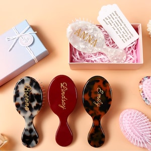 Può includere: Una collezione di spazzole per capelli ovali con nomi personalizzati in corsivo dorato. I design includono tartarugato bianco, bordeaux tinta unita e tartarugato marrone. Una spazzola è in una scatola regalo rosa con un biglietto bianco.