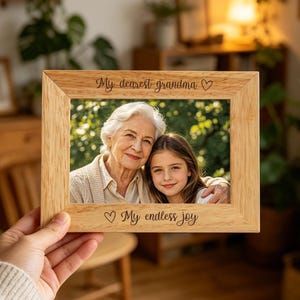 Puede incluir: Un marco de fotos rectangular de madera clara con texto grabado. El borde superior dice My dearest grandma con un icono de corazón, y el borde inferior dice My endless joy con un icono de corazón. El marco contiene una foto de una mujer mayor y una niña.
