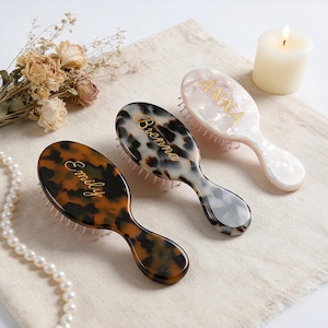 May include: Three oval hairbrushes with handles, each featuring a distinct pattern and a gold-colored name printed on the back. One brush has a dark brown tortoiseshell pattern labeled Emily, one has a grey tortoiseshell pattern labeled Brenna, and one has a white pearlescent pattern labeled Anna.