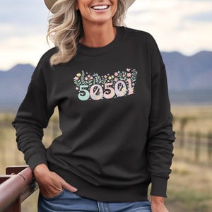Peut inclure: Sweat-shirt noir avec le numéro "50501" aux couleurs pastel et des motifs floraux. Le design comprend des teintes bleu clair, rose et vert. Le sweat-shirt est de style col rond.