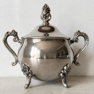 May include: A polished silver-toned sugar bowl with a rounded body, two ornate curved handles, and three decorative feet. The bowl features a matching removable lid topped with a detailed floral finial, reflecting light across its smooth surface.