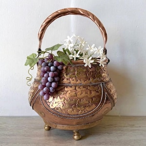 May include: A copper-colored metal basket with a curved handle and three small feet. The exterior features a repeating embossed pattern and is filled with a cluster of artificial purple grapes with green leaves and a bouquet of small white flowers.