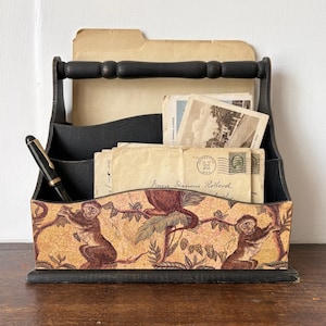 May include: Black wooden desk organizer with a handle, holding stationery and documents. The front features a decorative panel with monkeys and foliage in shades of brown and yellow. A black pen rests in a side compartment.