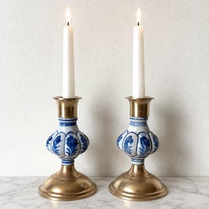May include: A pair of matching candle holders featuring a brass base and top, with a central ceramic section decorated in a blue floral pattern. Each holder supports a tall, slender white taper candle with a lit flame, creating a balanced display.