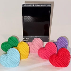 May include: A collection of colorful, heart-shaped objects with a knitted texture. The hearts are in various colors, including green, yellow, blue, pink, red, and purple. A black Instax Fujifilm box is behind the hearts.