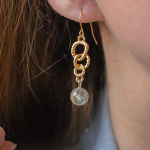 May include: Gold-coloured dangle earrings. The earrings feature a series of linked circles leading to a clear orb containing a delicate, white, feathery design. The earrings are shown worn on an ear.