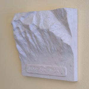 Könnte beinhalten: Eine weiße, strukturierte Reliefkarte einer Berglandschaft. Die rechteckige Karte trägt unten den Text "Jackson Hole, Wyoming". Die Karte ist an einer hellgelben Wand befestigt.