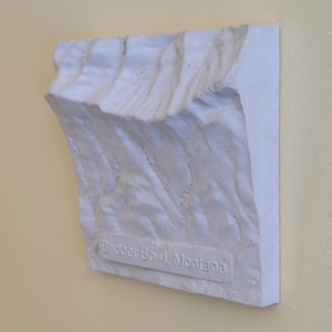 Może przedstawiać: Białe, teksturowane dzieło sztuki ściennej przedstawiające pasmo górskie. Element jest prostokątny z zakrzywioną górą i zawiera tekst "Bridger Bowl, Montana" na dole. Grafika jest zamontowana na jasnożółtej ścianie.