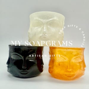 May include: Three decorative candle holders in white, black, and orange. Each holder features a face design with multiple faces. The text "MY SOAPGRAMS" and "ARTISAN GIFTS" are visible. The holders are likely for candles or small plants.