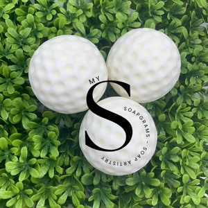 May include: Three white golf ball shaped soaps with a textured surface. The soaps are on a bed of green artificial grass. The text "MY SOAPGRAMS - SOAP ARTISTRY" is visible on one of the soaps.