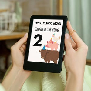 May include: A black e-reader displays a birthday graphic with the text "OINK, CLUCK, MOO! TAYLOR IS TURNING 2." The graphic features a brown cow, a pink pig, and a white chicken, all wearing party hats. The e-reader is held by a person.