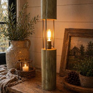 May include: A wooden floor lamp with a cylindrical design. The lamp has a warm, glowing bulb within a metal frame, with wooden sections at the top and bottom. The lamp is on a wooden surface, creating a cosy, inviting atmosphere. The lamp's height is approximately 76 cm.