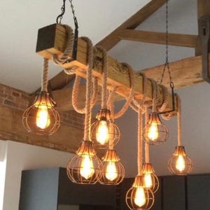 May include: A wooden beam chandelier with rope detailing. Seven caged light bulbs hang from the beam, providing a warm light. The fixture is suspended from the ceiling by chains, creating a unique lighting design. The overall design is rustic.