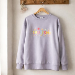 May include: A lavender crewneck sweatshirt with the word "AUTISM" embroidered in colorful letters. The word is accompanied by sun, cloud, and star designs. Below the word is the phrase "acceptance, understanding, joy". The sweatshirt is hanging on a wooden rack.