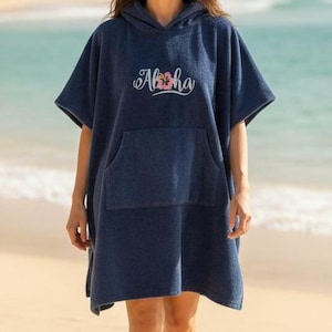 May include: A navy blue hooded beach poncho with a front pocket. The word "Aloha" is embroidered on the chest in a cursive font with a pink flower accent. The poncho is made of a soft, absorbent material, perfect for the beach or pool.