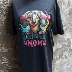 May include: Black t-shirt featuring a colorful illustration of a dachshund with the text "Dachshund MOM" in pink. The dog illustration has vibrant colors, including blue, pink, and yellow. Paw prints are included in the design.