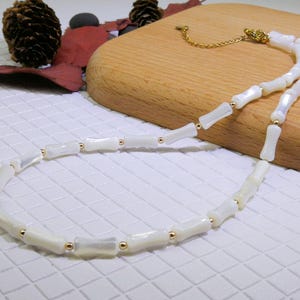 Puede incluir: Un collar blanco con un diseño similar al bambú. El collar presenta segmentos rectangulares con detalles dorados. El collar está sobre una superficie blanca con un fondo de madera.