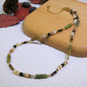 Puede incluir: Un collar de cuentas con una mezcla de cuentas verdes, marrones, negras y blancas. El collar tiene una cadena y un cierre de color dorado. Las cuentas son de varias formas y tamaños, incluyendo cilíndricas y redondas. El collar se exhibe sobre una superficie de madera.