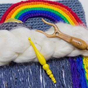 May include: A white yarn cloud with a rainbow crocheted wall hanging, a yellow crochet hook, and a wooden yarn swift.