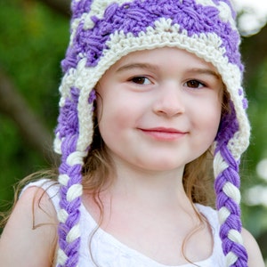 May include: A young girl wearing a purple and white crocheted hat with long braids. The hat has a crocheted pattern and is tied at the top with a white tassel.