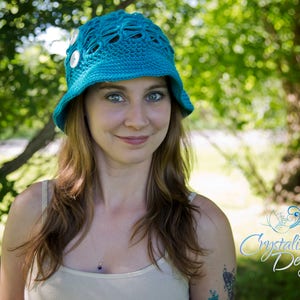 May include: A woman wearing a teal blue crocheted sun hat with white buttons. The hat has a wide brim and a lacy design. The woman has long brown hair and is wearing a white tank top.