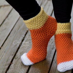 Puede incluir: Un par de calcetines de ganchillo hechos a mano con una combinación de colores de caramelo de maíz. Los calcetines tienen una puntera blanca, un cuerpo naranja y un puño amarillo. Los calcetines se muestran sobre una superficie de madera.