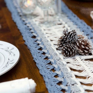 Op de afbeelding: Een blauw en wit gehaakte tafelloper met een gefranjerde rand, weergegeven op een donkere houten tafel. Drie dennenappels liggen op de loper. Witte borden en een theekopje zijn links zichtbaar.