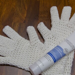 May include: Two white crocheted gloves and a tube of hand cream labeled "Moisture Therapy Intensive Healing & Repair" on a wooden surface.